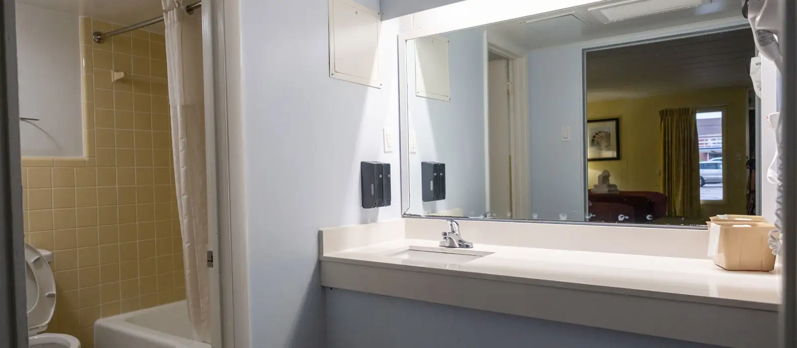 A bathroom with a toilet, a shower with a white curtain, and a vanity with a sink and mirror reflecting a bedroom and outside view