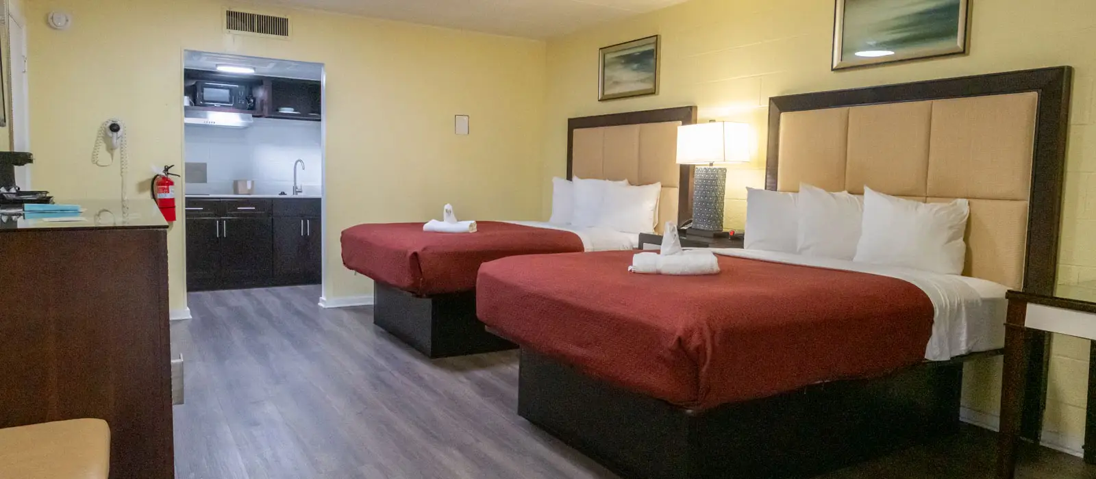 A guest room with two beds with red bedspreads, a bedside table with a lamp, and a kitchenette with a microwave and sink