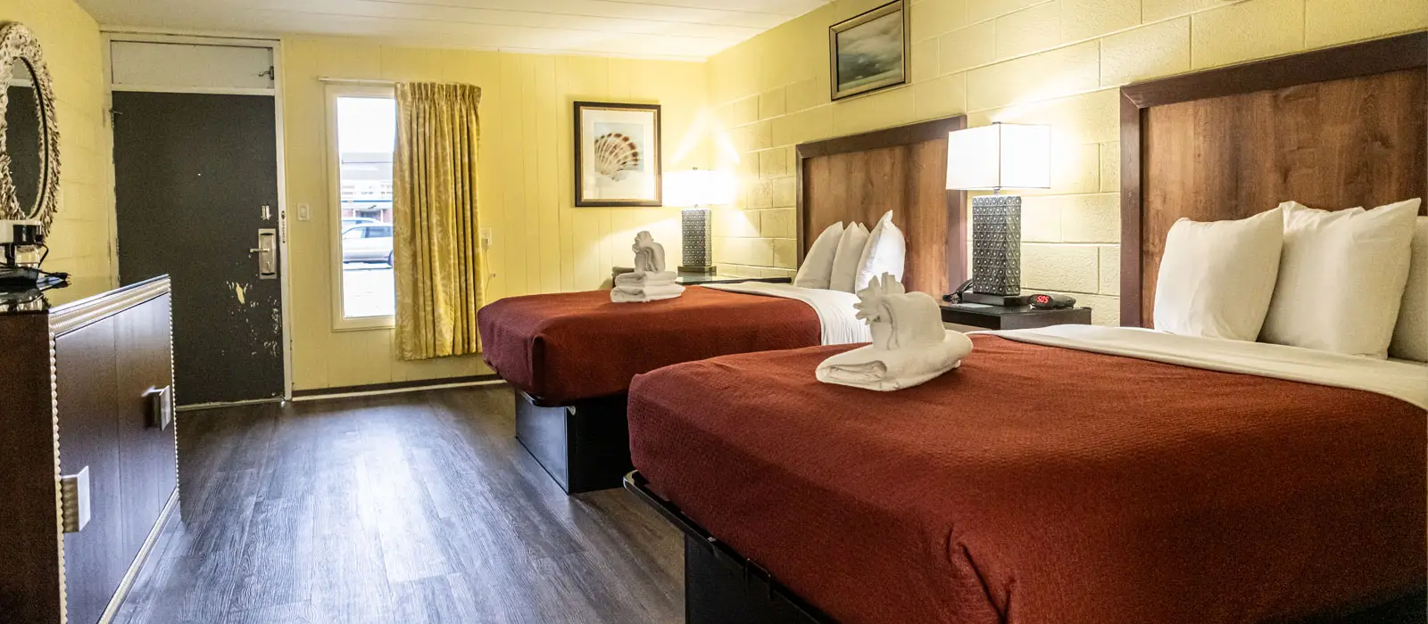 A guest room with two beds with red bedspreads, a dresser with a mirror, and a window with yellow curtains