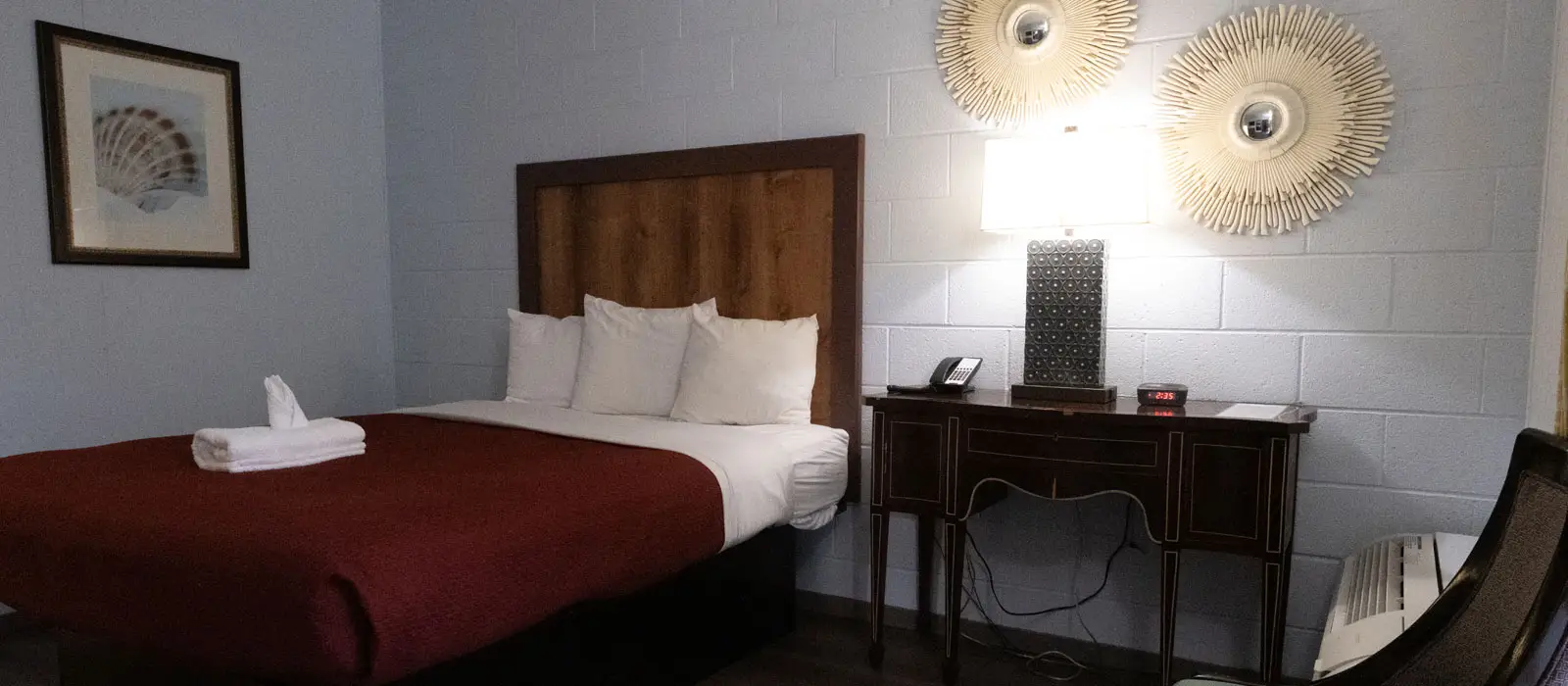 A guest room with a bed dressed in white and red bedding, a wooden headboard, a framed seashell picture, and a dark wood side table with a lamp and telephone