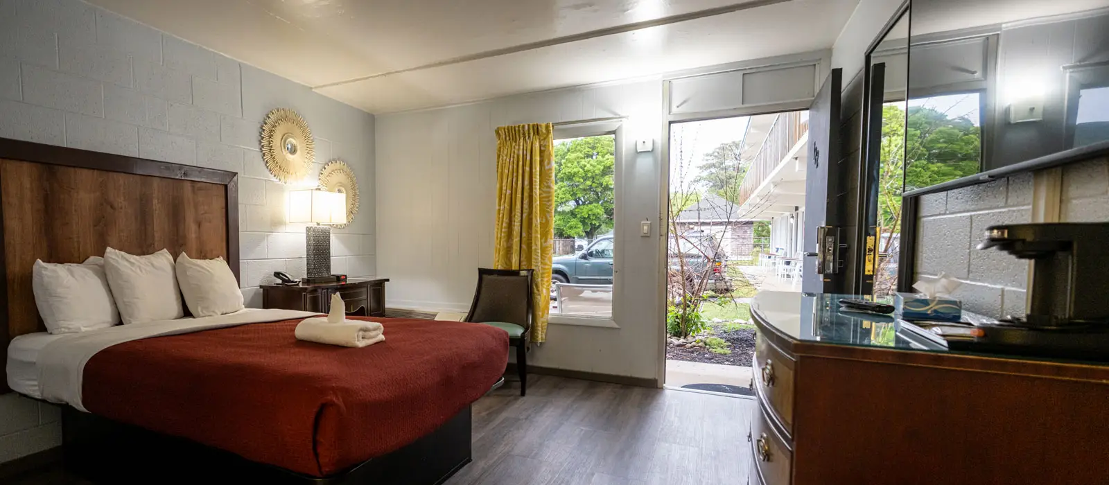 A hotel room with a bed with a red bedspread, a bedside table with a lamp, a chair, and an open door leading outside