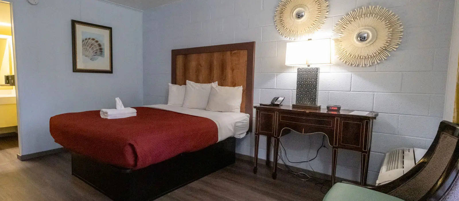 A guest room with a bed with a red duvet, a wooden headboard, a dark wood desk with a lamp, and two round wall decorations