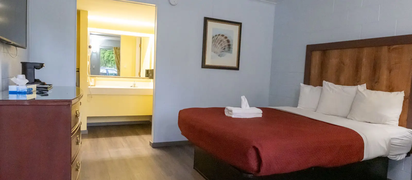 A guest room with a bed with a red duvet, a wooden headboard, a dresser with a TV and coffee maker, and a bathroom with a vanity