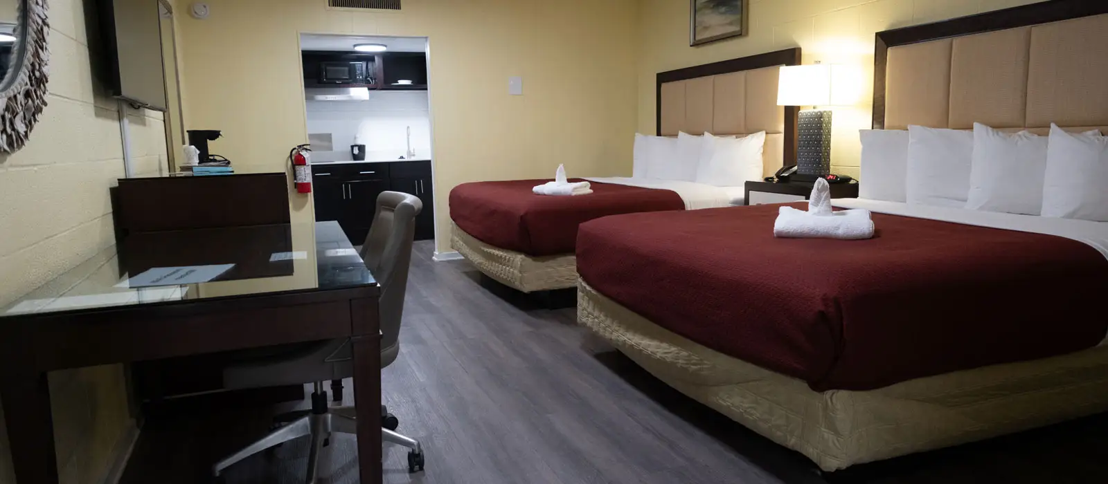 A guest room with two beds, a desk and chair, and a kitchenette visible through an open doorway