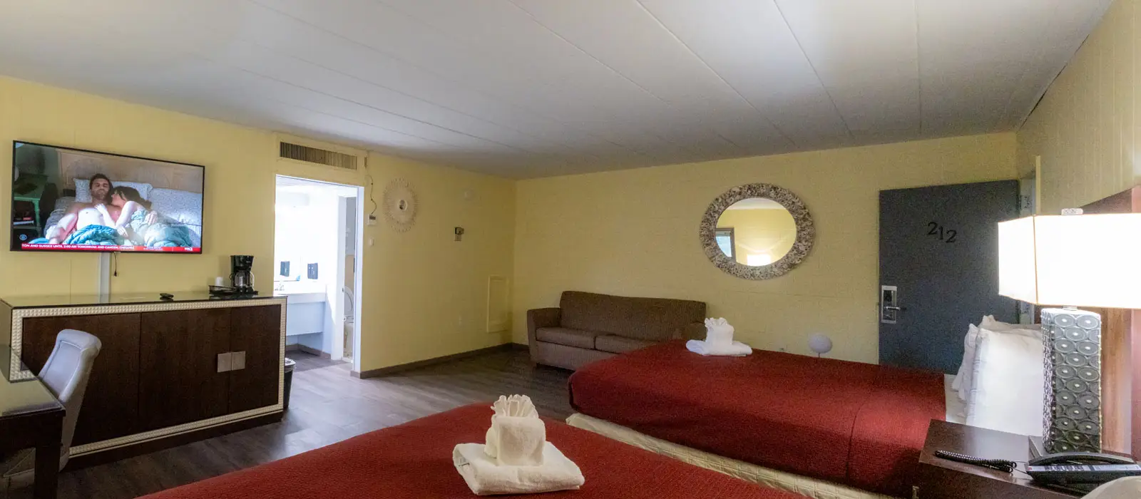 A hotel room with two beds with red bedspreads, a wall-mounted TV, a dark wood cabinet, a sofa and a round mirror
