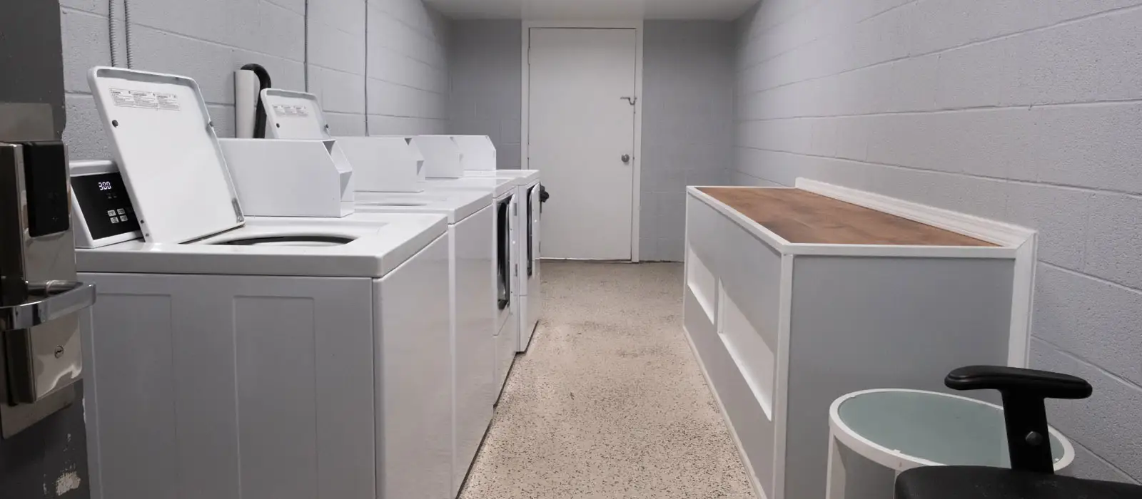 A laundry room with several white washing machines and dryers, a long wooden folding counter, and a white door at the far end