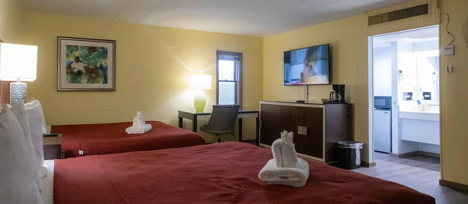 A hotel room with two beds, a desk, a wall-mounted TV, and an open doorway to a bathroom