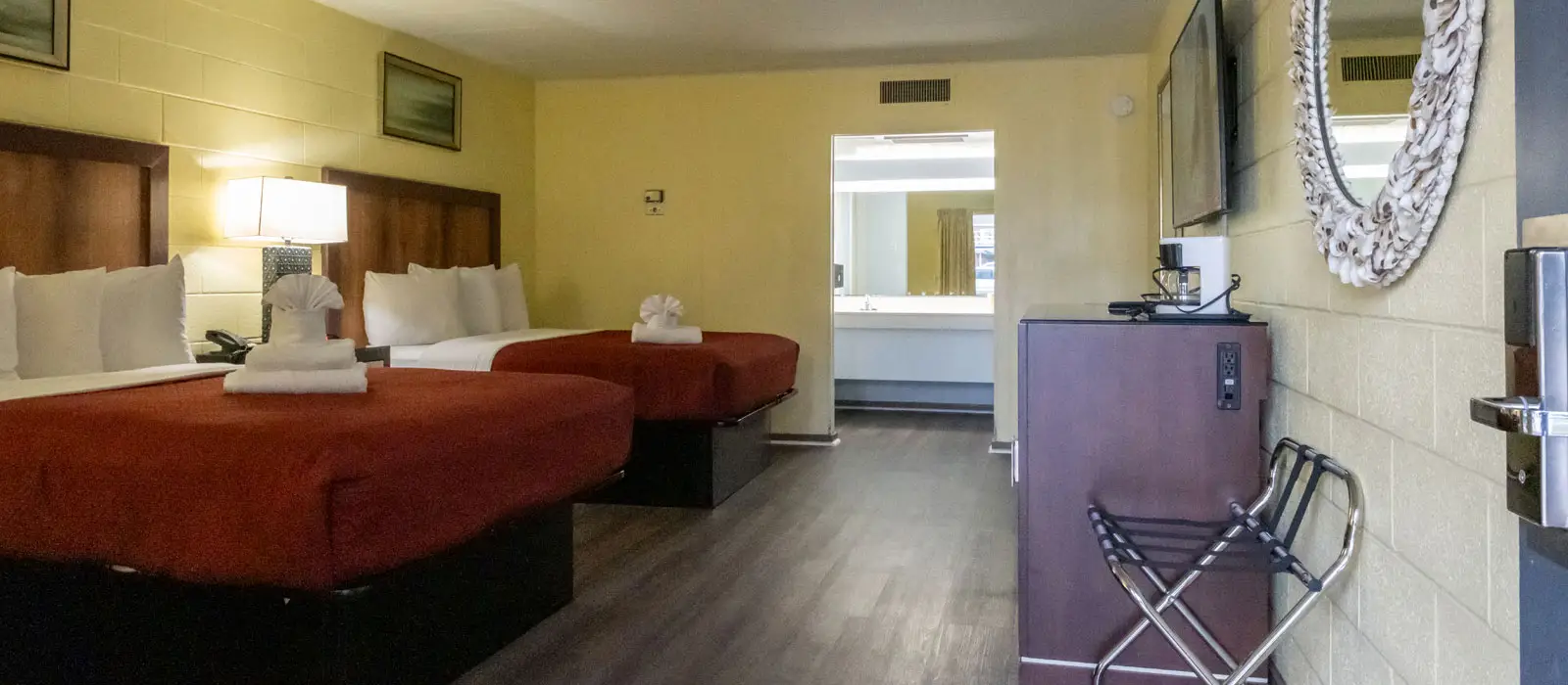A guest room with two beds with red bedding, a wall-mounted TV, a mirror and a luggage rack