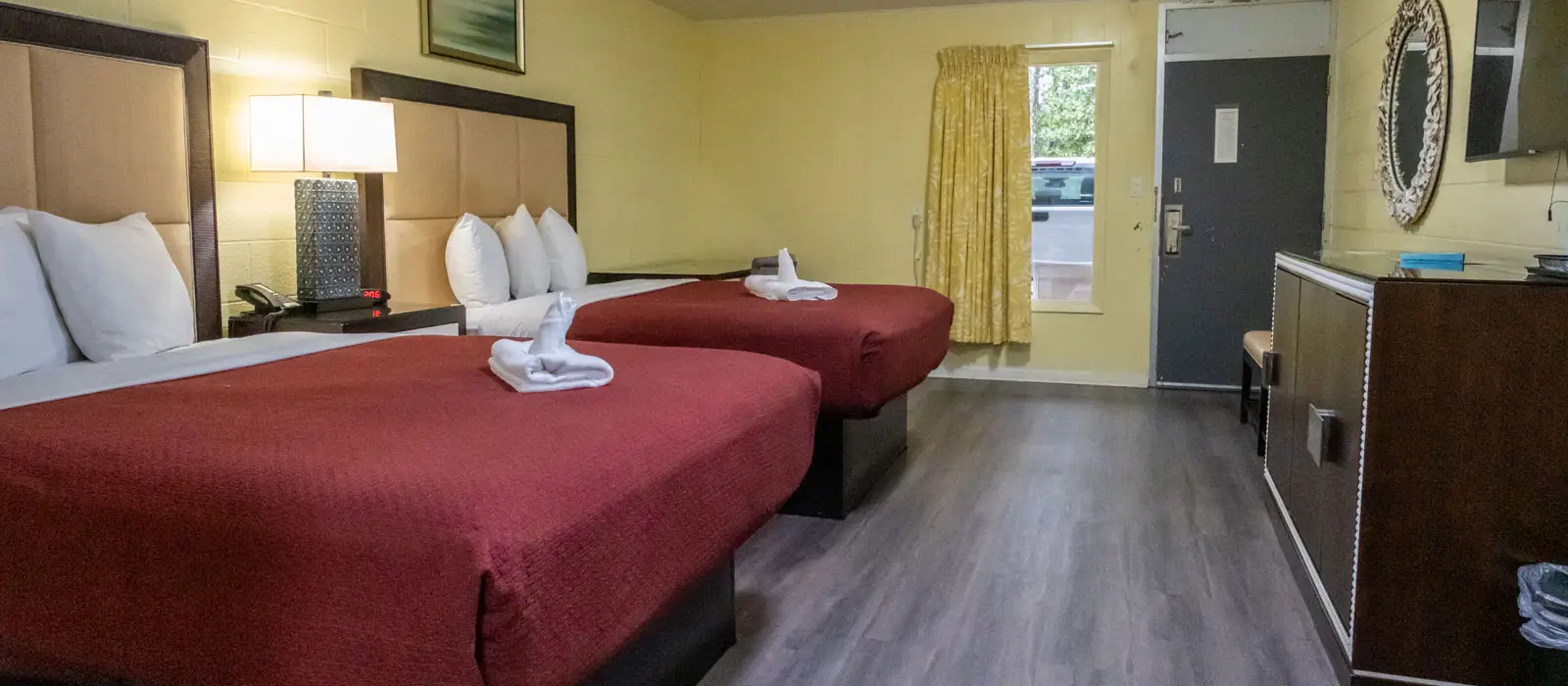 A guest room with two beds, a bedside table with a lamp and telephone, a window with yellow curtains, and a dresser with a mirror and television