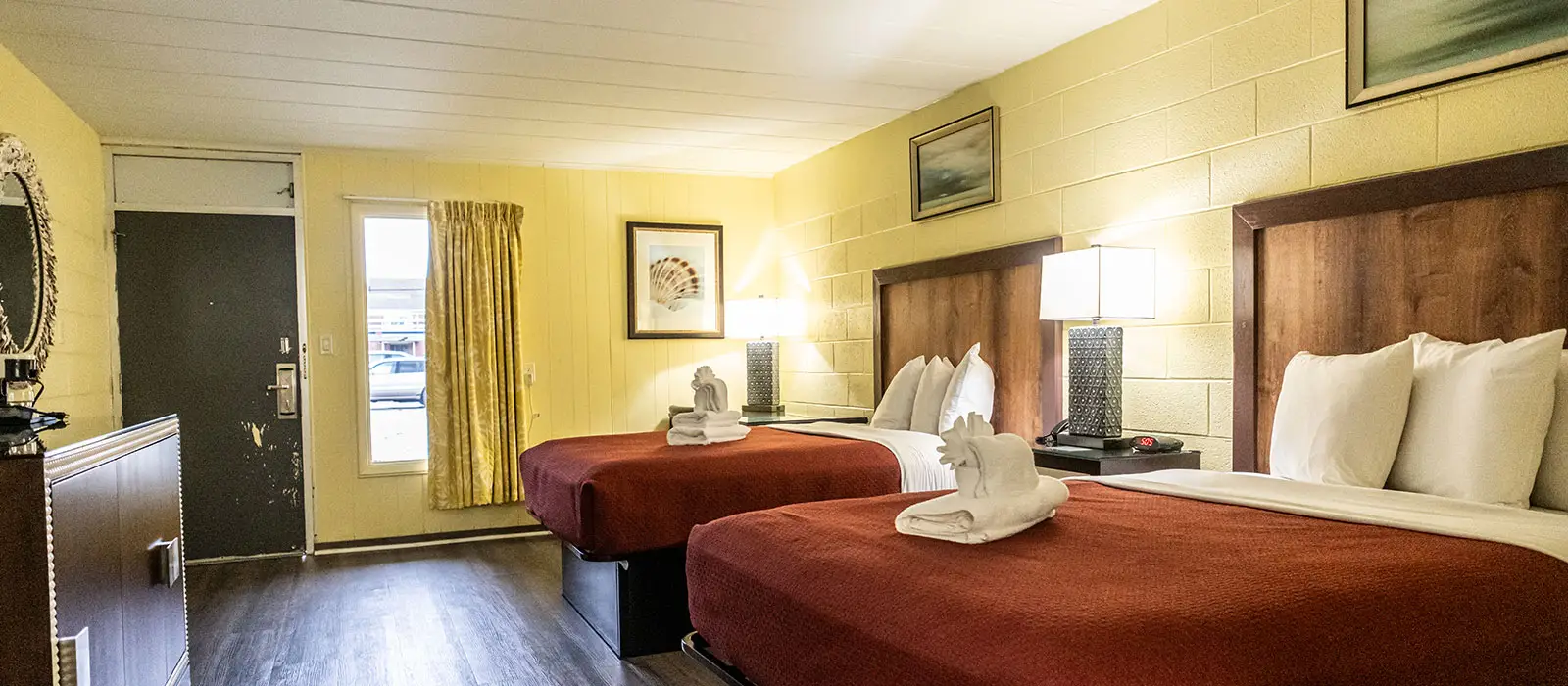 A hotel room with two beds, each with a red bedspread and white towels, and a window with a view of a street