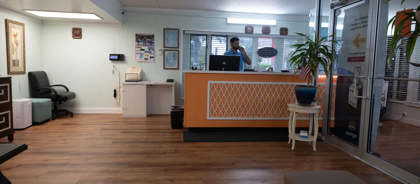 A hotel lobby with a man on the phone behind an orange reception desk, a waiting area with a chair and a small table, and a glass door