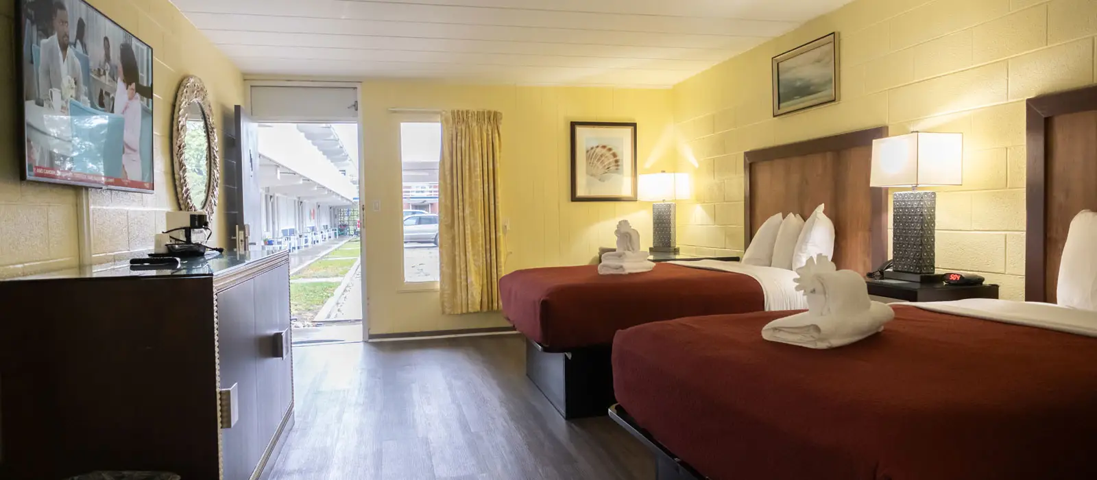 A hotel room with two beds with red bedspreads, a wall-mounted television, a dresser and an open door leading to an outdoor walkway