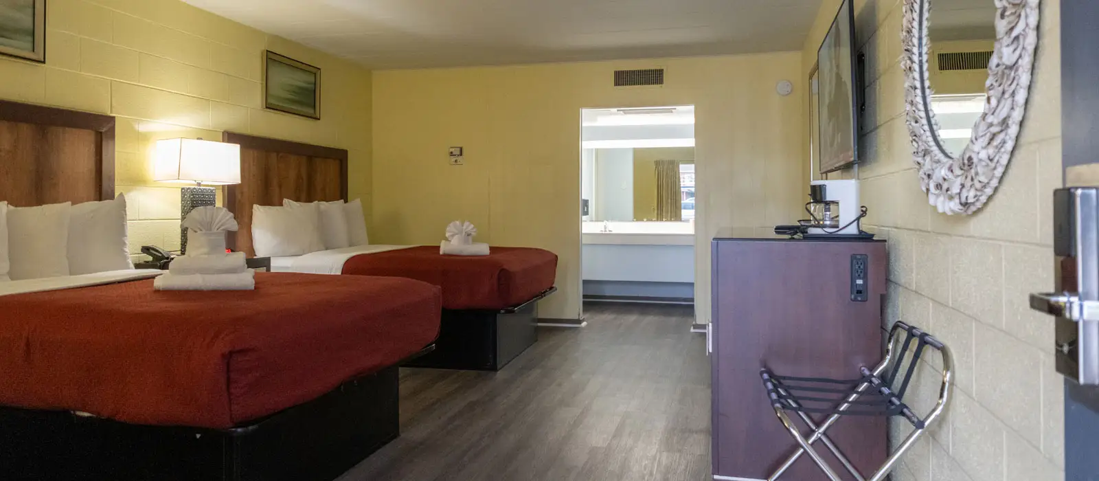 A hotel room with two beds, a bedside table with a lamp and telephone, and a bathroom vanity visible through an open doorway