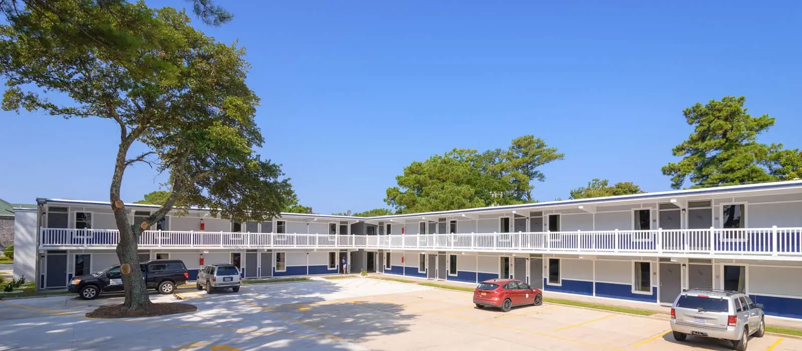 A two-storey motel with grey walls, blue accents and white railings, with cars parked in the lot and trees around the building