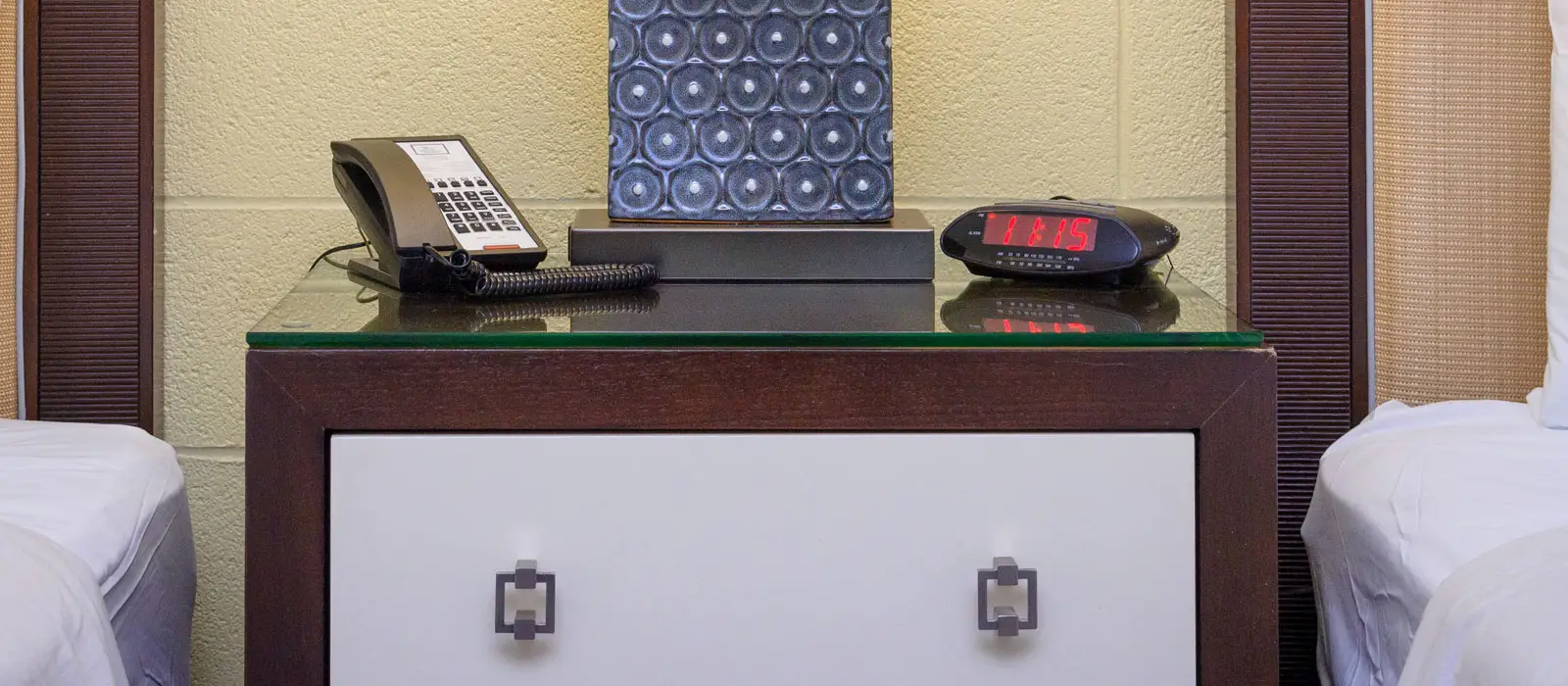 A bedside table with a telephone, a decorative lamp and a digital alarm clock displaying 8:18