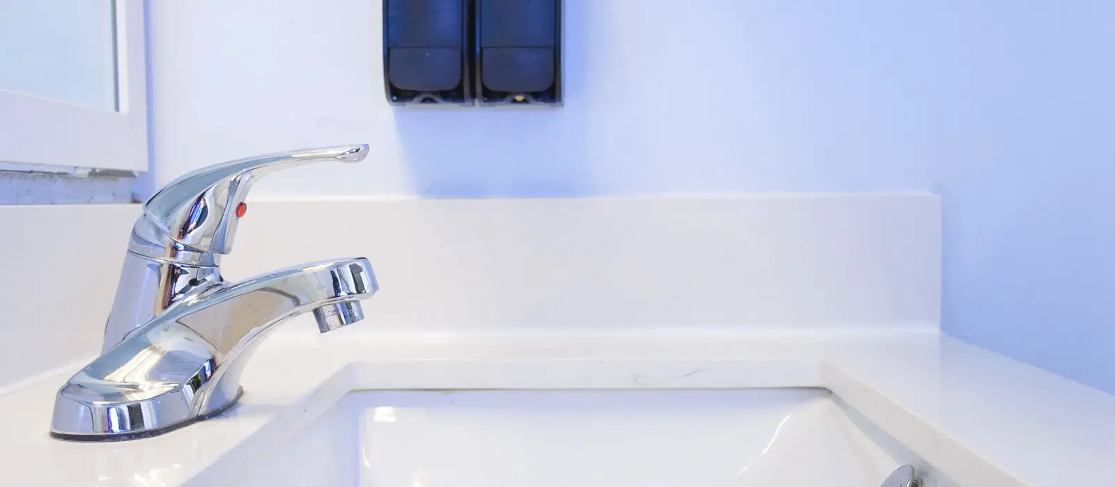 A chrome tap with a red indicator button is mounted on a white countertop with a sink, below two black wall-mounted dispensers