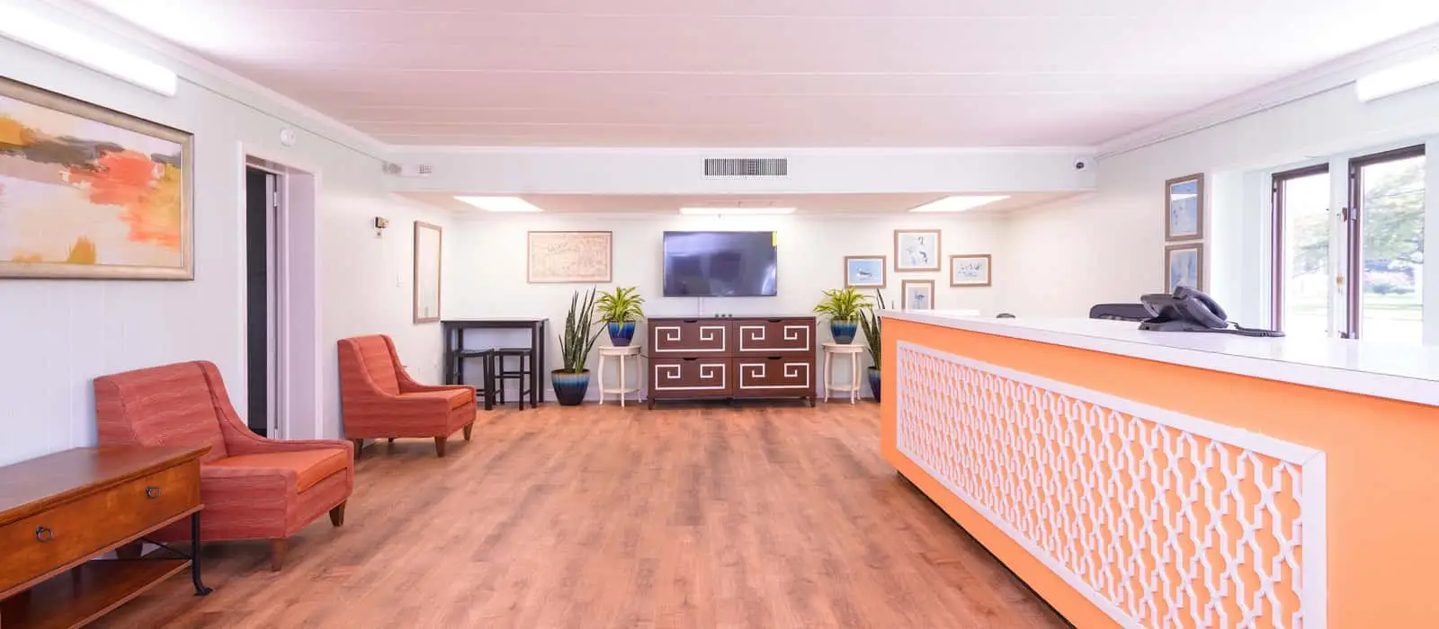 A hotel lobby with a long orange and white reception desk, two orange armchairs, and a TV mounted above a dark wood cabinet