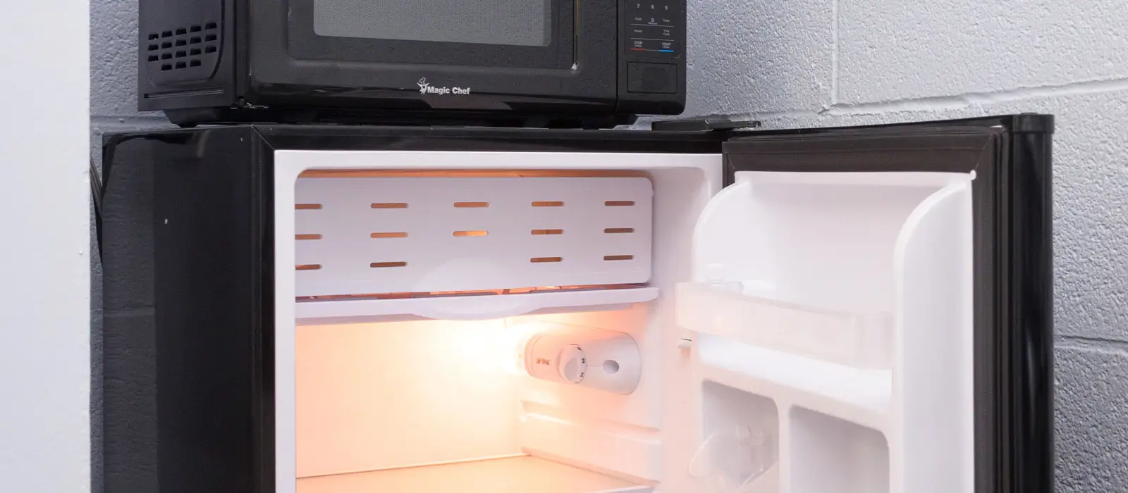 A black microwave sits atop a black mini-fridge with its door open, revealing a light-up interior
