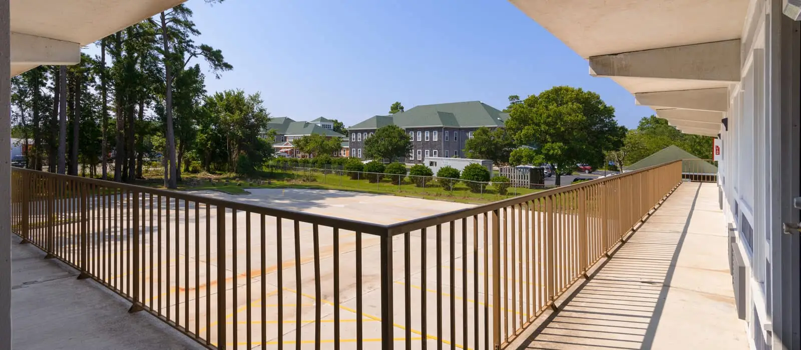 A long outdoor walkway with a brown railing, looking out over a parking area and several buildings with green roofs