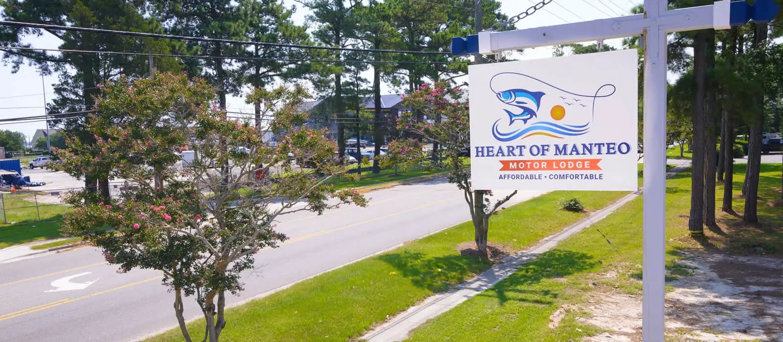 A white sign with a blue fish logo and the words "HEART OF MANTEO MOTOR LODGE AFFORDABLE COMFORTABLE" stands beside a road with trees and buildings in the background