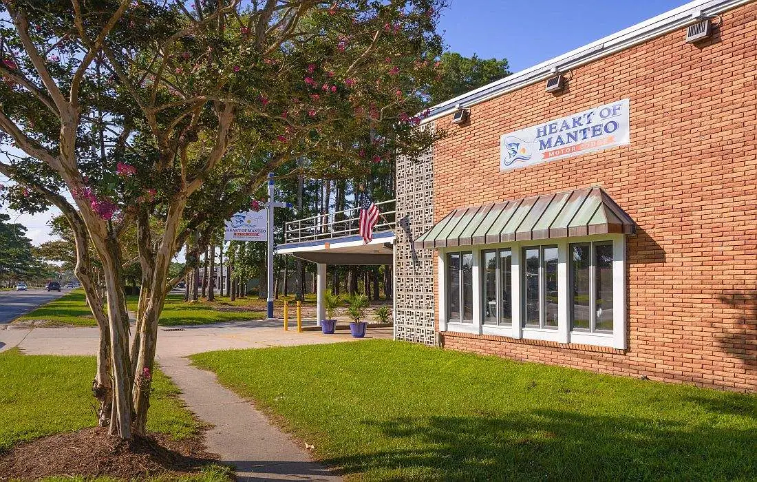 A brick building with a sign reading "HEART OF MANTEO MOTOR LODGE" above a window with an awning, next to a tree-lined street