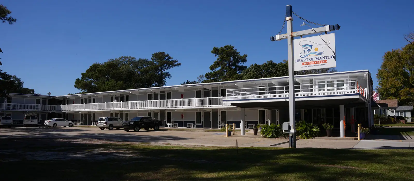 A two-storey white building with blue accents and white railings, with a sign reading "Heart of Manteo Motor Lodge" on the right