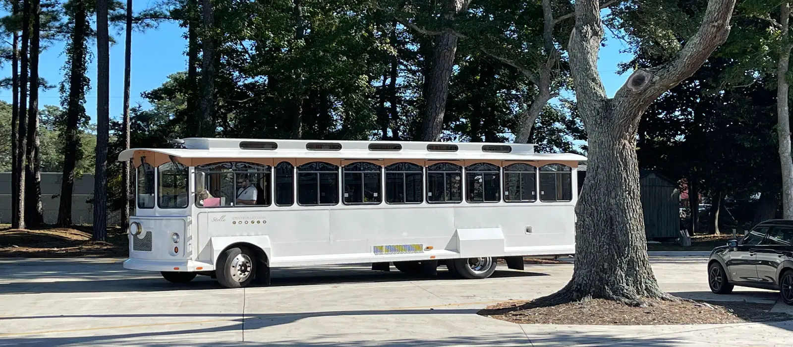 A white trolley bus with passengers is parked on a paved area next to several trees, with a dark car visible on the right