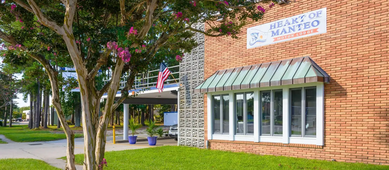 A red brick building with a sign that reads "Heart of Manteo Motor Lodge" above a window with a green awning, and a tree with pink flowers in the foreground