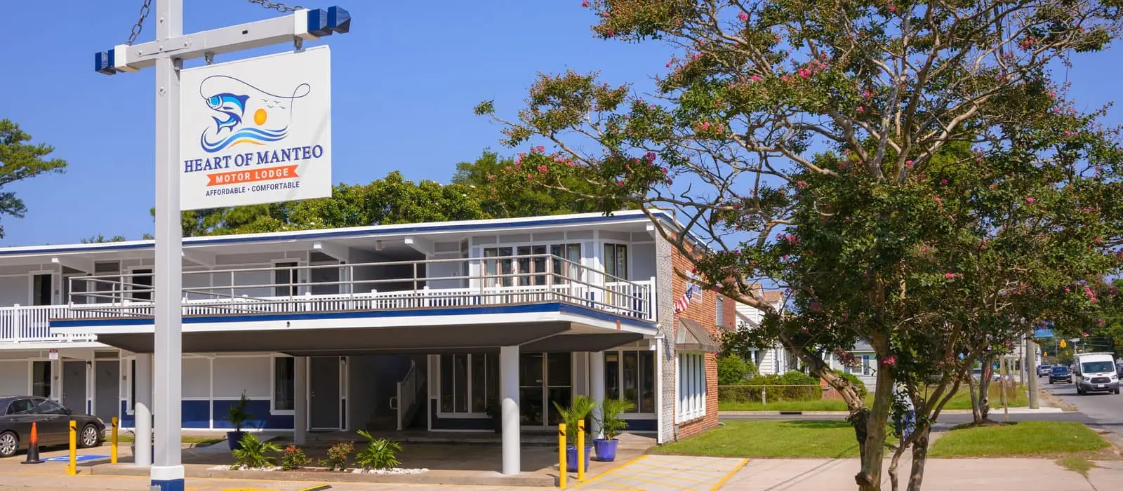 The exterior of a two-storey motor lodge with a white and blue sign that reads "Heart of Manteo Motor Lodge" and a street with cars